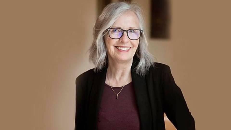 Sharon Thesen, a woman with shoulder-length white hair and glasses, wears a plum coloured top and a black blazer and smiles at the camera.