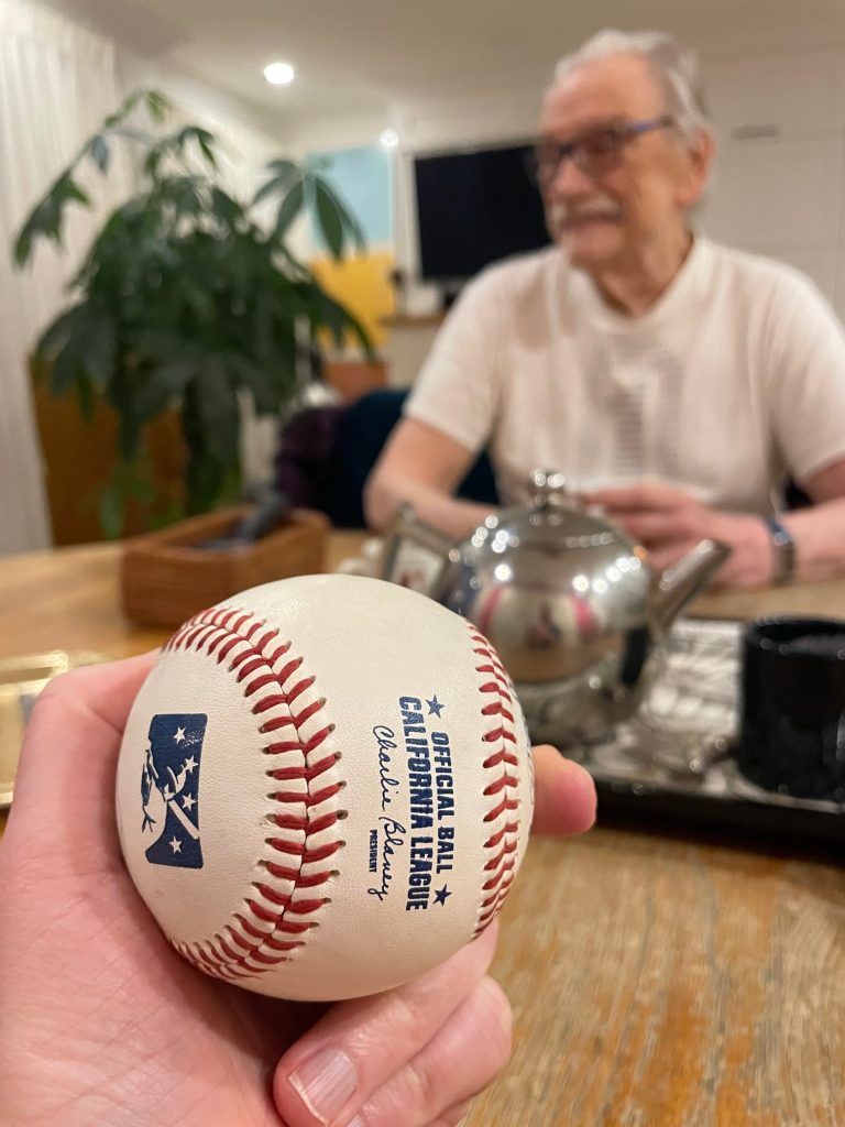 A hand holds a California League Official Ball. An out-of-focus George Bowering smiles in the background.